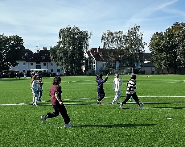 Eine Gruppe von Menschen spielt auf einem grünen Kunstrasenplatz Fußball. Im Hintergrund sind Bäume und ein Gebäude zu erkennen.