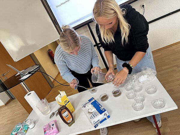 Zwei weibliche Auszubildende beuge sich über einen Tisch, auf dem Glasschälchen, Feuchttücher, Honig, Küchentücher, Wasser etc., stehen und rühren Heilerde an.