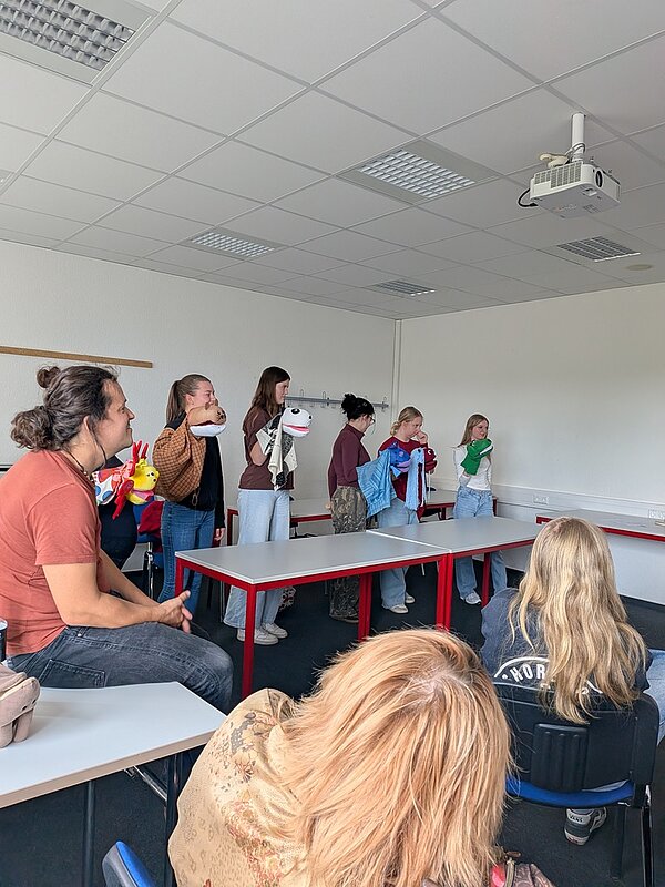 Eine Gruppe junger Azubis steht in einem hellen Raum hinter Tischen und führt mit ihren selbst gebastelten bunten Handpuppen ein kleines Theaterstück im Rahmen des Puppen-Workshops auf.