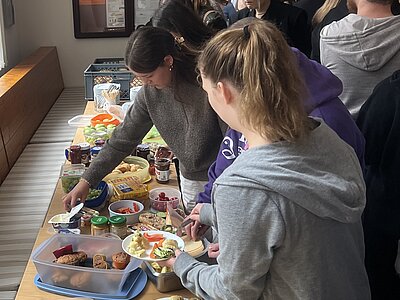 Eine Reihe von Menschen steht an einem Buffet-Tisch und bedient sich an verschiedenen Speisen. Es gibt eine Auswahl an Lebensmitteln, darunter Gemüse, Brote, Aufstriche und Snacks. Die Menschen unterhalten sich und füllen ihre Teller.
