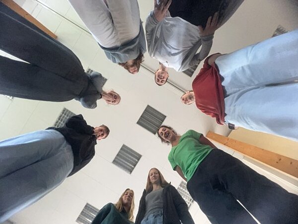 Gutgelaunter gemeinsamer Start ins Schuljahr Junge Erwachsene stehen zusammen in einem Kreis. Die Perspektive ist von unten nach oben. Dadurch erscheinen die Personen sehr langgezogen. Die Personen blicken in die Kamera und lächeln.