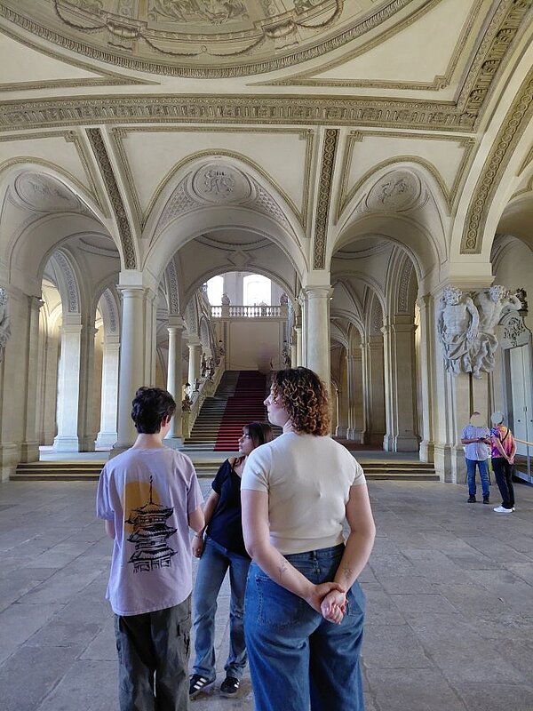nnenansicht der Würzburger Residenz: Eine Gruppe von Personen steht in einem prunkvollen, barocken Saal mit hohen Gewölbedecken, verzierten Säulen und einem roten Teppich auf der Treppe. Die Architektur zeigt typische Merkmale des Rokoko mit reichhaltigem Stuck und Ornamenten.