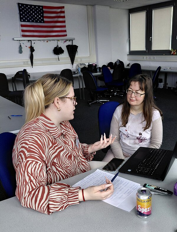 Eine Frau erklärt einer anderen Frau etwas, vor ihr auf dem Tisch liegen Zettel, Stift und Laptop; im Hintergrund ist ein Klassenzimmer mit einer US-amerikanischen Flagge an der Wand zu sehen.