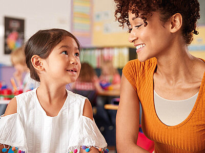 Auf dem Bild sind zwei Personen im Klassenzimmer zu sehen. Die Person links trägt eine weiße Bluse mit bunten Pompons an den Ärmeln, während die Person rechts ein orangefarbenes Oberteil und lockiges Haar hat. Im Hintergrund sind Kinderzeichnungen, pädagogische Poster und andere Lernmaterialien an den Wänden zu erkennen.