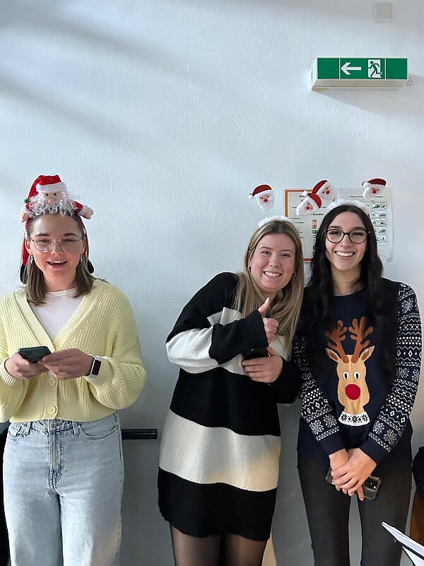 Drei junge Frauen stehen nebeneinander, tragen weihnachtliche Accessoires und lächeln in die Kamera, eine zeigt den Daumen nach oben.