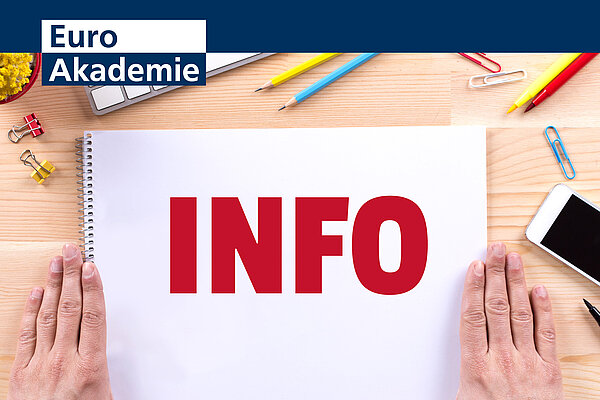 Die Aufschrift "Info" in roten Lettern steht auf einem Zeichenblock geschrieben, der auf einem Holztisch liegt und neben dem rechts und links die Hände einer Person flach aufliegen. Um den Block verteilt liegen Stifte, Büroklammern und ein Handy. Links oben befindet sich das Logo der Euro Akademie.