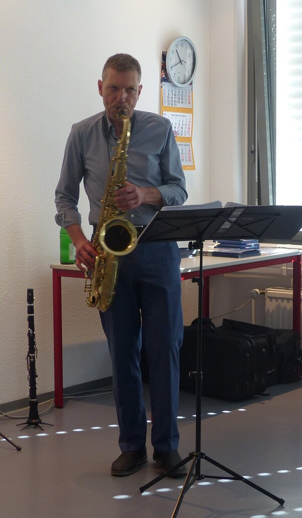 Ein Mann in einem hellblauen Hemd und einer Jeans spielt Saxophone. Vor ihm steht ein Notenständer und hinter ihm ist eine weiße Wand und ein Tisch, neben dem eine Klarinette steht.