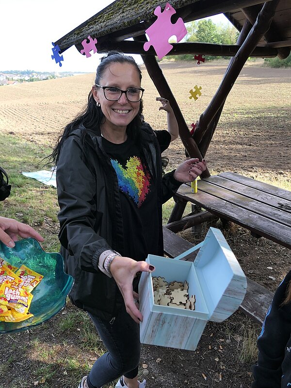 Frau mit dunkler Brille und langen Haaren steht unter einem hölzernen Unterstand auf einer Wiese. Sie trägt eine schwarze Jacke und ein schwarzes T-Shirt mit einem bunten Herzmotiv aus Puzzleteilen. In ihrer Hand hält sie eine offene, hellblaue Box mit Puzzleteilen. Über ihr hängen große, pinkfarbene Puzzleteile am Dach des Unterstands. Neben ihr reicht eine Person eine Schale mit Süßigkeiten. Im Hintergrund ist ein Feld und eine Bank aus Holz zu sehen.