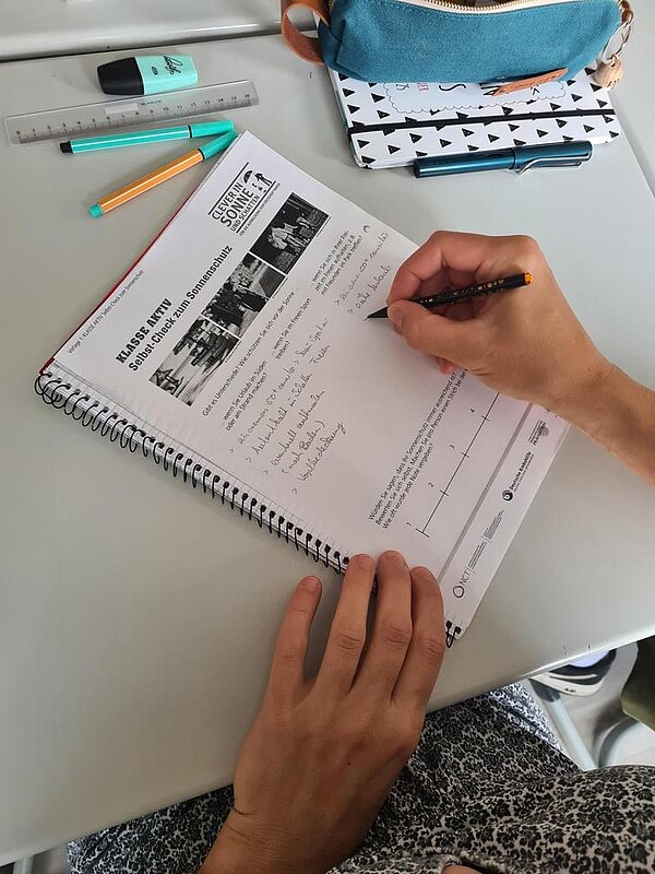 Auszeichnung „Clever in Sonne und Schatten“ 2024 Ein Schüler macht sich Notizen in einem Handout. Blick auf die Hände und den Block, daneben liegen verschiedene Stifte und ein Lineal.