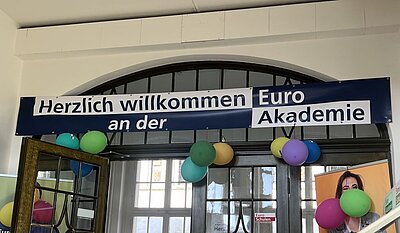Ein Willkommensbanner mit der Aufschrift „Herzlich willkommen an der Euro Akademie“ hängt über einem Eingang. Darunter sind bunte Luftballons befestigt, die eine festliche Stimmung zum Schulstart vermitteln.