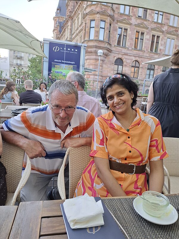 Zwei Personen sitzen lächelnd an einem Tisch im Außenbereich eines Restaurants vor historischer Gebäudekulisse.