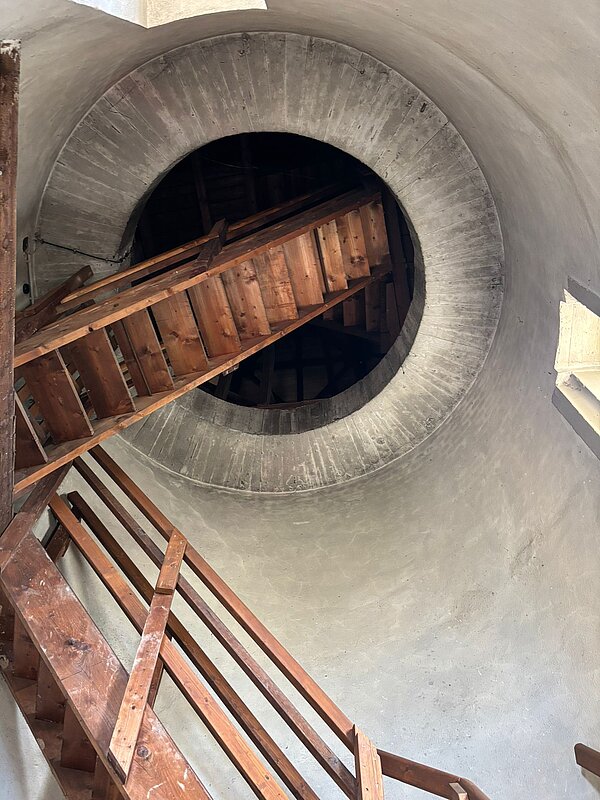 Holztreppe in einem engen, runden Treppenturm mit Betonwänden, die nach oben zu einer weiteren Öffnung führen.