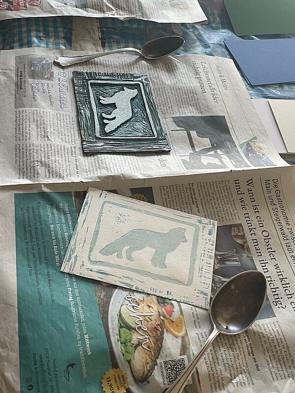 Zwei kleine Druckplatten mit Tiermotiven liegen neben Metalllöffeln auf ausgebreiteten Zeitungen nach einem kreativen Druckprojekt.