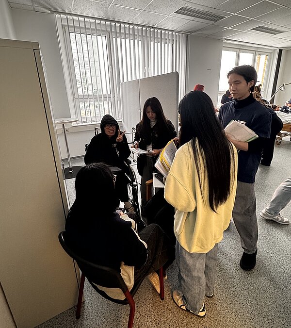 Fünf Auszubildende der Euro Akademie Bitterfeld-Wolfen stehen und sitzen in einem Raum mit großen Fenstern und vertikalen Jalousien. Zwei Azubis sitzen auf Stühlen, drei stehen um sie herum. Ein Azubi hält ein aufgeschlagenes Buch, eine andere hält einen Block und einen Stift. Im Hintergrund sind ein Schrank, ein Whiteboard und weitere Möbelstücke zu sehen.