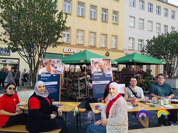 Im Vordergrund sitzen mehrere Menschen an Holztischen auf einem öffentlichen Platz. Die Tische sind mit bunten Papierbastelarbeiten und Regenbogenmotiven dekoriert. Im Hintergrund stehen zwei große Infotafeln mit Text und Bildern, die auf ein Angebot oder eine Veranstaltung hinweisen. Dahinter befinden sich grüne Marktstände mit Blumen und Pflanzen sowie mehrstöckige Gebäude mit hellen Fassaden. Die Szene wirkt freundlich und einladend.