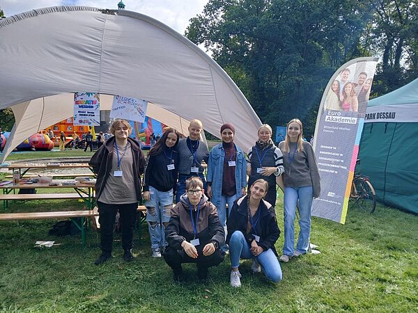 Eine Gruppe junger Menschen steht und kniet lächelnd vor einem weißen Zelt auf einer grünen Wiese. Im Hintergrund sind bunte Attraktionen und ein Banner mit der Aufschrift "Euro Akademie" zu sehen.