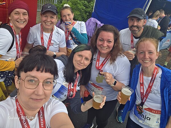 Selfie von zehn Teammitgliedern mit Medaillen und Getränken. Alle strahlen und zeigen ihre Medaillen, die Stimmung ist ausgelassen und feierlich.