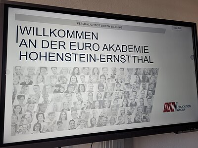 Auf einem großen Bildschirm ist eine Präsentation zu sehen mit den Worten "Willkommen an der Euro Akademie Hohenstein-Ernstthal" und dem Logo der ESO Education Group.