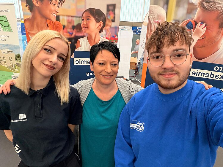 Drei Personen stehen dicht beieinander vor Infowänden der Euro Akademie Altenburg und lächeln in die Kamera; die beiden äußeren tragen Kleidung mit Euro-Akademie-Logo.