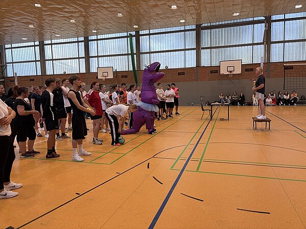 In einer Sporthalle stehen mehrere Menschen nebeneinander auf dem gelben Hallenboden. Eine Person trägt ein auffälliges, lila Dinosaurierkostüm mit weißem Tutu. Die Gruppe blickt zu einer weiteren Person, die auf einem Stuhl steht und spricht. Im Hintergrund sitzen Zuschauer auf Bänken vor einer Backsteinwand mit großen Fenstern.