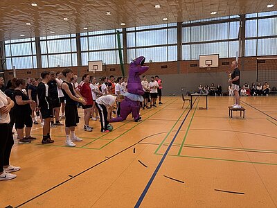 In einer Sporthalle stehen mehrere Menschen nebeneinander auf dem gelben Hallenboden. Eine Person trägt ein auffälliges, lila Dinosaurierkostüm mit weißem Tutu. Die Gruppe blickt zu einer weiteren Person, die auf einem Stuhl steht und spricht. Im Hintergrund sitzen Zuschauer auf Bänken vor einer Backsteinwand mit großen Fenstern.