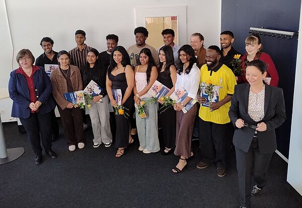 Eine Gruppe von 17 Personen steht in einem hellen Raum mit blauer Wand und weißer Tür. Viele halten Blumensträuße und Zertifikate oder Urkunden in den Händen. Die Gruppe wirkt feierlich und stolz, möglicherweise bei einer Abschlussfeier. Die Personen sind unterschiedlich gekleidet, einige tragen formelle Kleidung, andere lässigere Outfits.