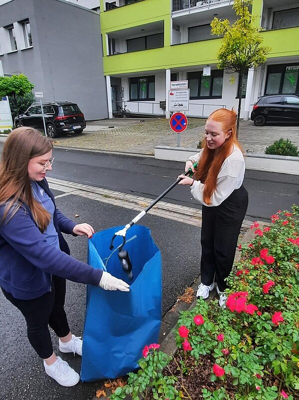 Tag der sauberen Umwelt Zwei Schülerin fischen mit einem Sammelstab eine Flasche aus einer Blumenrabatte mit Rosen und befördern sie in einen großen blauen Plastiksack.