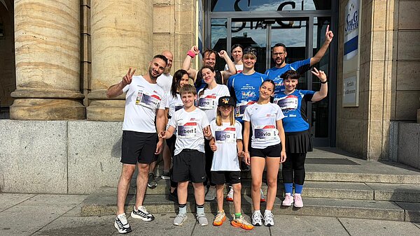 Eine Gruppe Menschen in Sporttrikots steht vor einem Sandsteingebäude. Die Menschen lächeln und strecken die Arme in die Höhe.