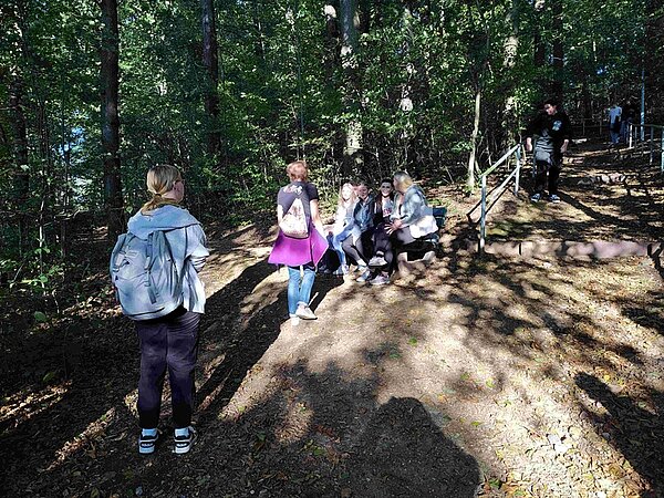 Schülerinnen sitzen auf einer Bank und Schüler gehen eine Treppe im Wald herunter. 