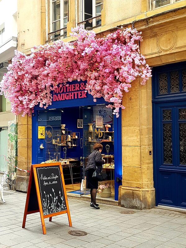 Schulfahrt nach Metz Blick auf das Schaufenster und Gebäude eines Schmuckgeschäftes namens Mother and Daughter. Das Gebäude ist gelb, die Fensterrahmen blau, vor der Tür steht eine Tafel, über dem Schaufenster hängen zahlreiche Kirschblüten und eine Dame bückt sich, um besser auf die Auslage im Schaufenster blicken zu können.