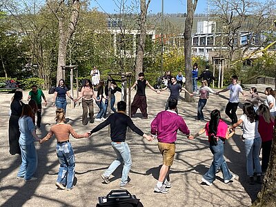 Eine große Gruppe von Menschen tanzt im Kreis unter freiem Himmel in einem Park; alle halten sich an den Händen, während andere im Hintergrund zusehen; die Szene wirkt fröhlich und gemeinschaftlich.