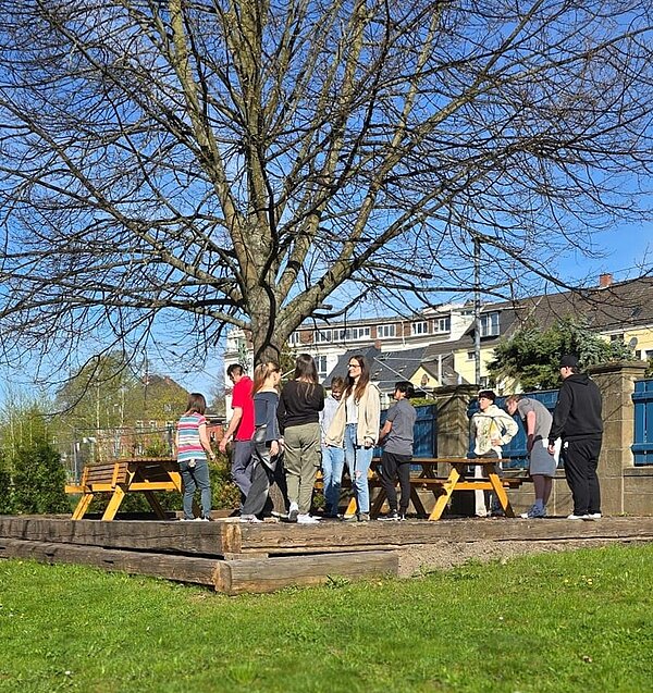 Foto der Schülerinnen und Schüler der Euro Akademie Hohenstein-Ernstthal stehen draußen in ihrem sogenannten Grünen Klassenzimmer, das sich auf einem Podest aus Holz und direkt unter einem großen, schönen Baum befindet, auf dem die Holztische und Bänke stehen.