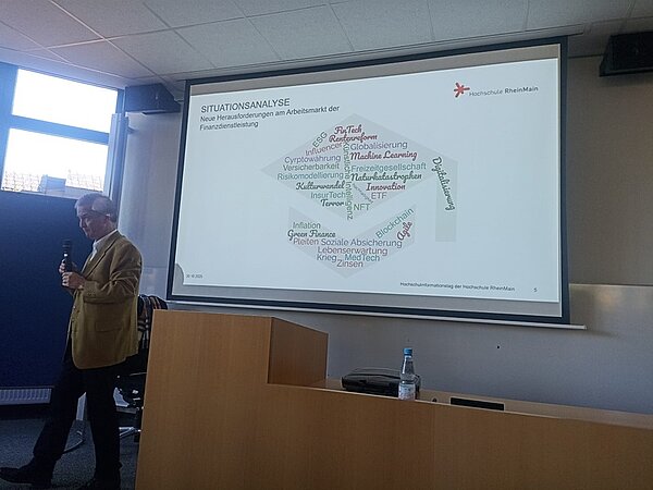 Blick auf die Präsentation auf der Leinwand zum Thema Situationsanalyse. Der Referent steht mit Mikrophon davor und hält seine Präsentation.