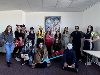 Eine Gruppe von jungen Menschen in kreativen und fantasievollen Kostümen posiert gemeinsam in einem Klassenzimmer. Zu sehen sind verschiedene Verkleidungen, darunter Disney-Figuren, Star-Wars-Charaktere, Feen und eine Person mit einer weißen Maske. Alle lächeln und präsentieren stolz ihre Outfits.