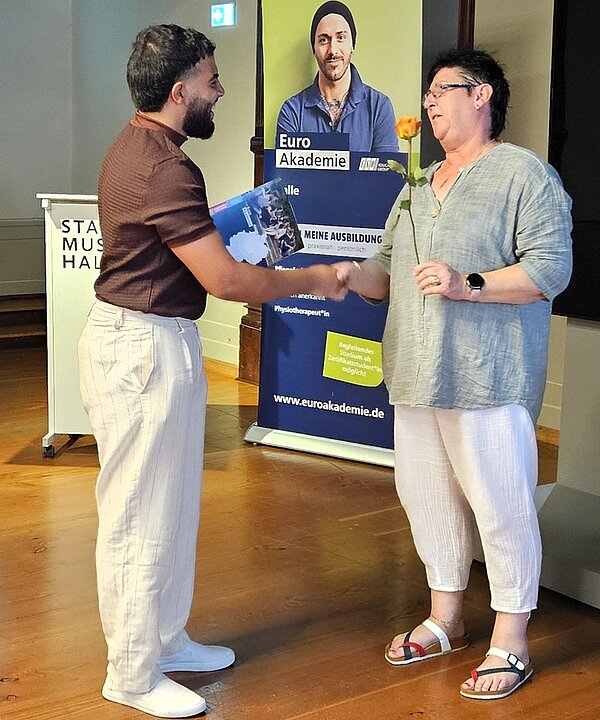 Lehrkraft überreicht einem Absolventen sein Abschlusszeugnis und eine Rose. 