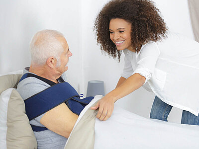 Eine lächelnde Pflegekraft hilft einem älteren Mann mit einer Armstütze, sich vom Bett aufzusetzen. Der ältere Mann trägt eine beige Hose und ein graues T-Shirt, die Pflegekraft trägt eine weiße Bluse und eine Jeans.
