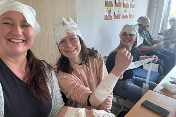 Zum Abschluss einen Erste-Hilfe-Kurs Im Vordergrund grinsen drei junge Mädchen in die Kamera und zeigen ihre Arme, die mit weißen Mullbinden umwickelt sind. Auch um ihre Köpfe haben sie Verbandsmaterial.