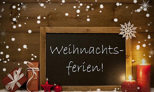 Ein winterliches Motiv mit einem Schiefertafel-Schild, auf dem ‚Weihnachtsferien!‘ steht. Im Hintergrund schneit es, daneben liegen ein Geschenk, ein roter Apfel, eine Kerze und ein Schneeflocken-Dekostern.