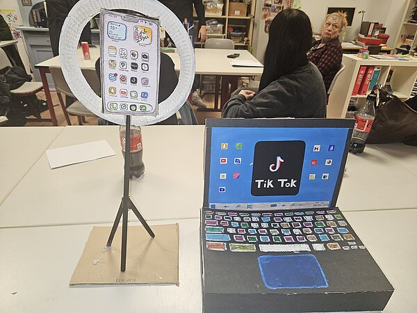 Ein aus Pappe gebautes Notebook steht neben einem Ständer mit Papier-Smartphone und einer Papier-Ringlampe.
