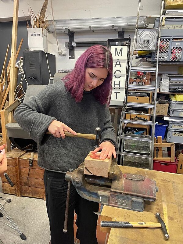 Eine Person mit violetten Haaren bearbeitet mit einem Holzhammer ein in einem Schraubstock auf einer Holzwerkbank eingespanntes Werkstück. Im Hintergrund sind Regale mit Werkzeug und Materialien zu sehen.