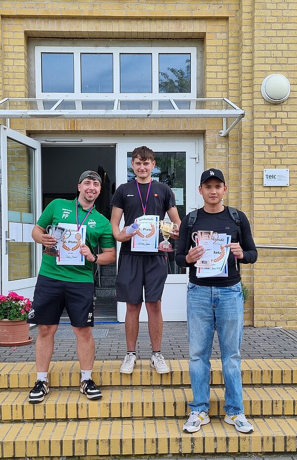Wikinger-Sportfest: Pflegeazubis erobern die nordische Arena Drei junge Männer stehen auf einer Treppe vor einem Gebäude. Sie halten Urkunden, Medaillen und Pokale und lächeln in die Kamera.