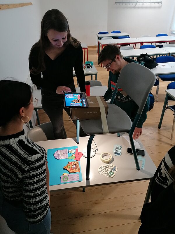 MeKoSax-Projekt „Trickfilmproduktion“ Schülerinnen und Schüler filmen mit einem an einem an einem Stuhl montierten Tablet ein blaues Papier mit Wolken ab.
