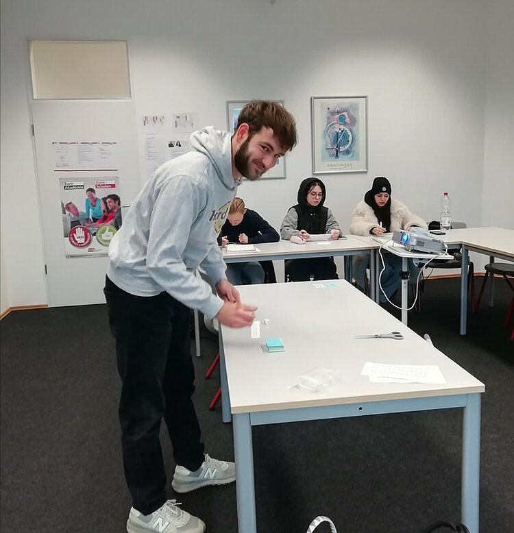 Mit Spaß im Spanisch Unterricht? – Bingo! Schüler steht lächelnd am Tisch im Klassenraum und wartet auf die richtigen Bingozahlen, wie seine Mitschüler*innen im Hintergrund.