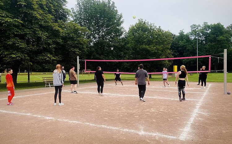 Sport frei! Eine Gruppe von jungen Menschen spielt auf einem Ascheplatz Volleyball. DAs Volleyballnetz ist auffällig pink.