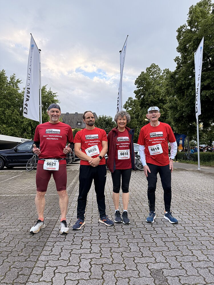 Vier Personen in roten Trikots der Euro Akademie und Sportschuhen stehen auf einem Parkplatz und lächeln in die Kamera.