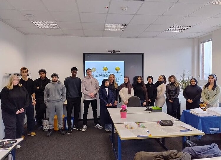 Mehrere Auszubildende stehen in einem Klassenraum in der Euro Akademie Berlin nebeneinander vor einem Bildschirm mit einer Willkommensnachricht und posieren für ein Erinnerungsfoto.