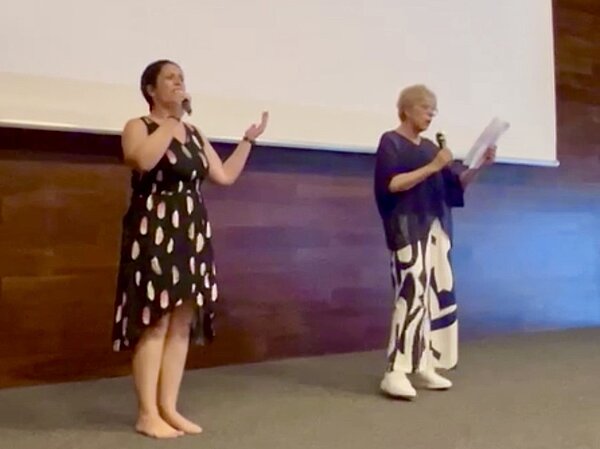 Zwei Frauen stehen auf einer Bühne und singen in Mikrophone. Eine hält dabei einen Zettel, die andere hält voller Inbrunst beim Singen die Hand hoch.