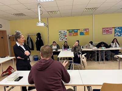 Eine Person in formeller Kleidung steht vorne in einem Klassenzimmer und spricht zu einer Gruppe junger Erwachsener, die an langen Tischen sitzen. Die Zuhörenden sind aufmerksam und blicken in Richtung der sprechenden Person. Einige von ihnen haben Hefte oder Stifte in der Hand. Im Hintergrund sind eine gelbe Wand mit bunten Plakaten, eine Tafel und ein Projektor zu sehen. Die Atmosphäre wirkt interaktiv und engagiert. Eine Person im Vordergrund sitzt mit dem Rücken zur Kamera und hört ebenfalls zu.