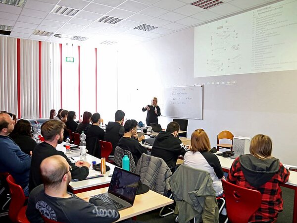 Eine Referentin hält eine Präsentation vor einer Gruppe von Zuhörern und Zuhörerinnen in einem Seminarraum. Die Teilnehmenden sitzen an Tischen mit Laptops und Notizbüchern, während der Referentin vorne steht und auf eine Projektionsleinwand zeigt.