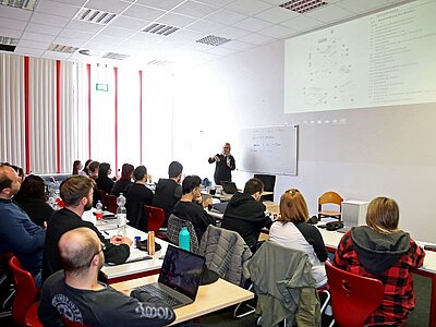  Eine Referentin hält eine Präsentation vor einer Gruppe von Zuhörern und Zuhörerinnen in einem Seminarraum. Die Teilnehmenden sitzen an Tischen mit Laptops und Notizbüchern, während der Referentin vorne steht und auf eine Projektionsleinwand zeigt.